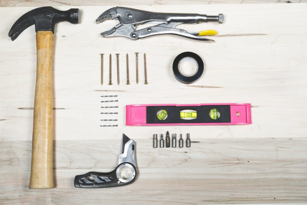 A neatly arranged collection of tools on a wooden surface includes a hammer, pliers, screws, nails, a roll of electrical tape, a pink level, a utility knife, and various screwdriver bits.