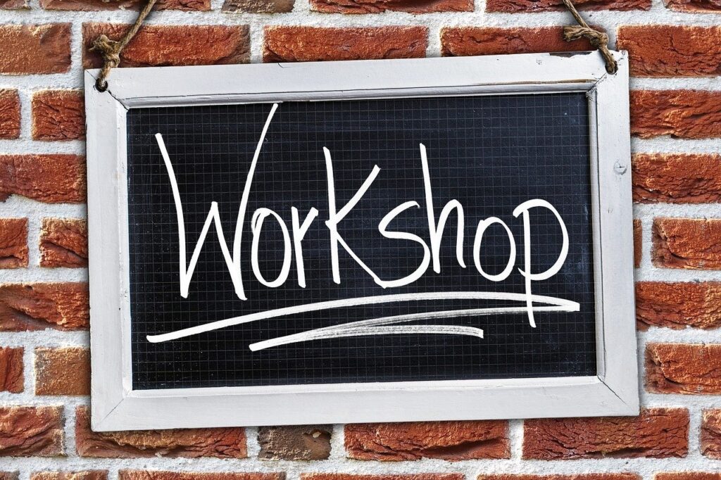 A black chalkboard with a white wooden frame hangs on a red brick wall. The word "Workshop" is written on the chalkboard in large, white cursive letters, with an underline below it.