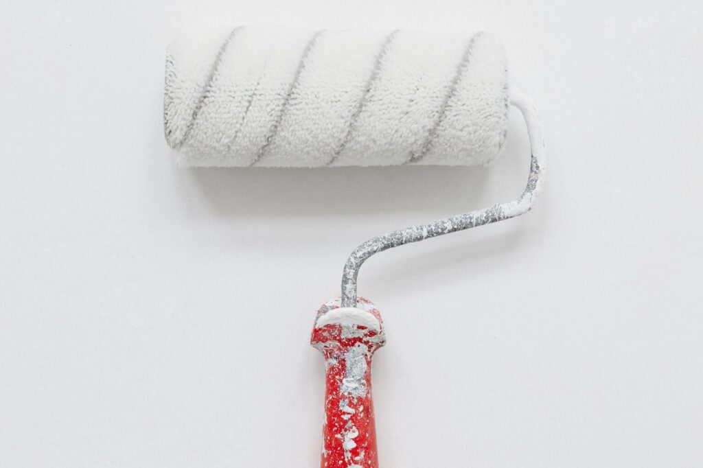 A paint roller with a white, slightly worn foam cover and red paint-splattered handle rests against a plain white background. The handle has a metallic extension connecting to the roller, which shows signs of paint use.