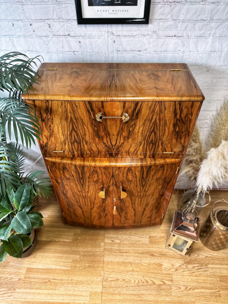 The Turnridge of London Cocktail Cabinet, a vintage 1950s walnut drinks unit with an Art Deco flair, stands against a white brick wall. It features a small upper compartment with a latch and two lower doors adorned with brass handles. Potted plants and a metal lantern are placed around it, enhancing the decor.