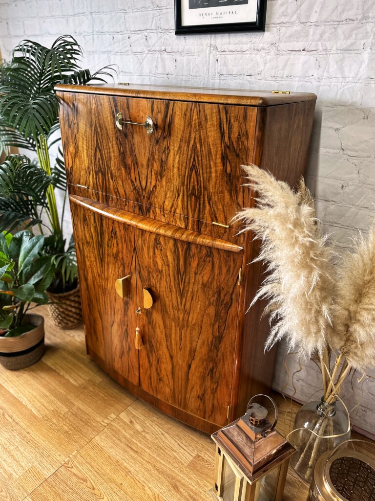 A Turnridge of London Cocktail Cabinet from the 1950s, featuring rich walnut with a swirl-patterned grain and brass handles, stands against a white brick wall. Various potted plants, including pampas grass, surround the vintage Art Deco drinks bar, creating a cozy and natural atmosphere in the room.