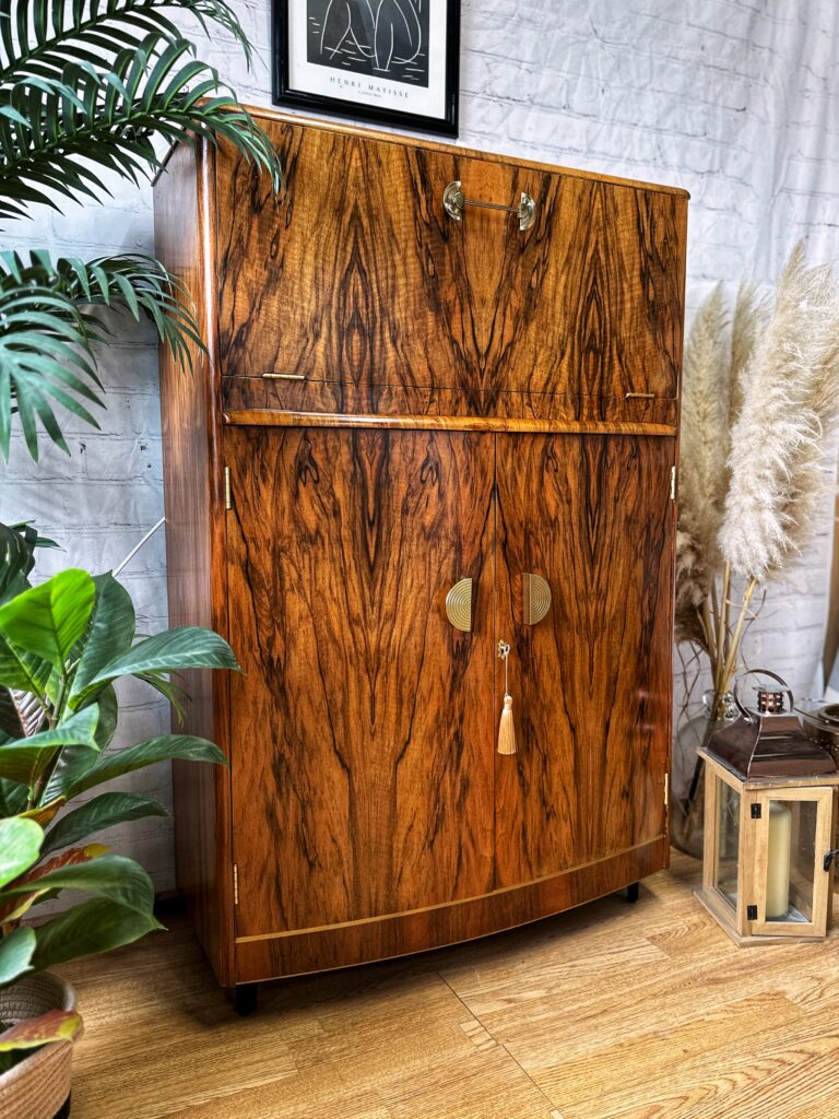 A Turnridge of London Cocktail Cabinet, exuding vintage Art Deco charm and crafted from walnut, stands against a white brick wall. The 1950s drinks unit features a drop-down door and two circular brass handles adorned with a tassel. It is surrounded by green potted plants and a lantern, with a framed print hanging above it.