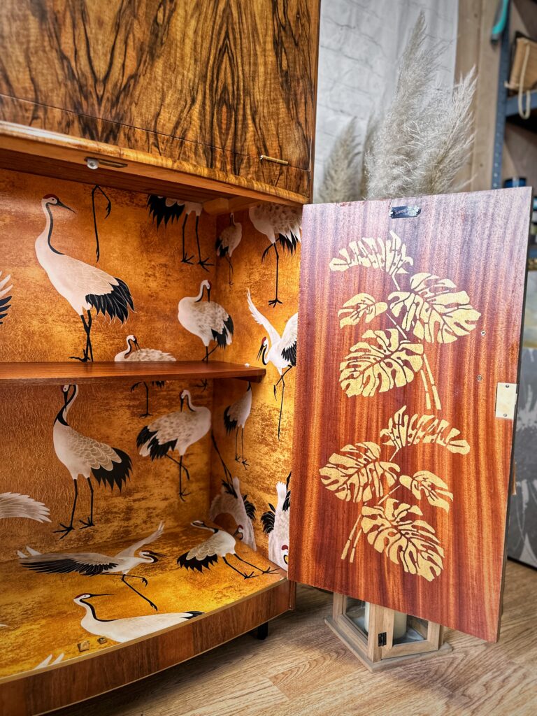 The Turnridge of London Cocktail Cabinet from the 1950s, crafted in walnut, is open to reveal two inner shelves adorned with an orange design featuring white cranes. The cabinet door, resting against the unit, showcases an intricate leaf pattern. The background features a brick wall and pampas grass in a vase.