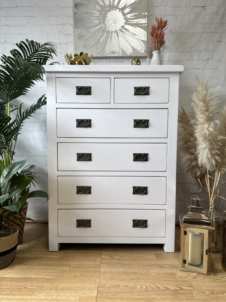 A white, six-drawer dresser with black handles stands against a white brick wall. On top, a tray with decor items and a round vase are displayed. Surrounding the dresser are potted plants, pampas grass, and a lantern on the floor. A floral artwork hangs above.