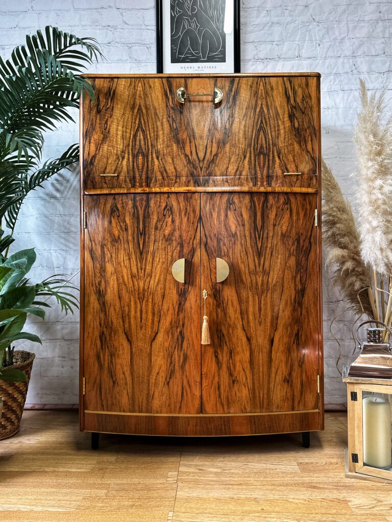 The Turnridge of London Cocktail Cabinet, a vintage drinks bar from the 1950s with distinct walnut grain patterns, stands against a light brick wall. It features two small upper doors and larger lower doors adorned with round gold handles and a tassel. The scene is elegantly decorated with plants and a lantern, while a framed abstract art piece hangs above.