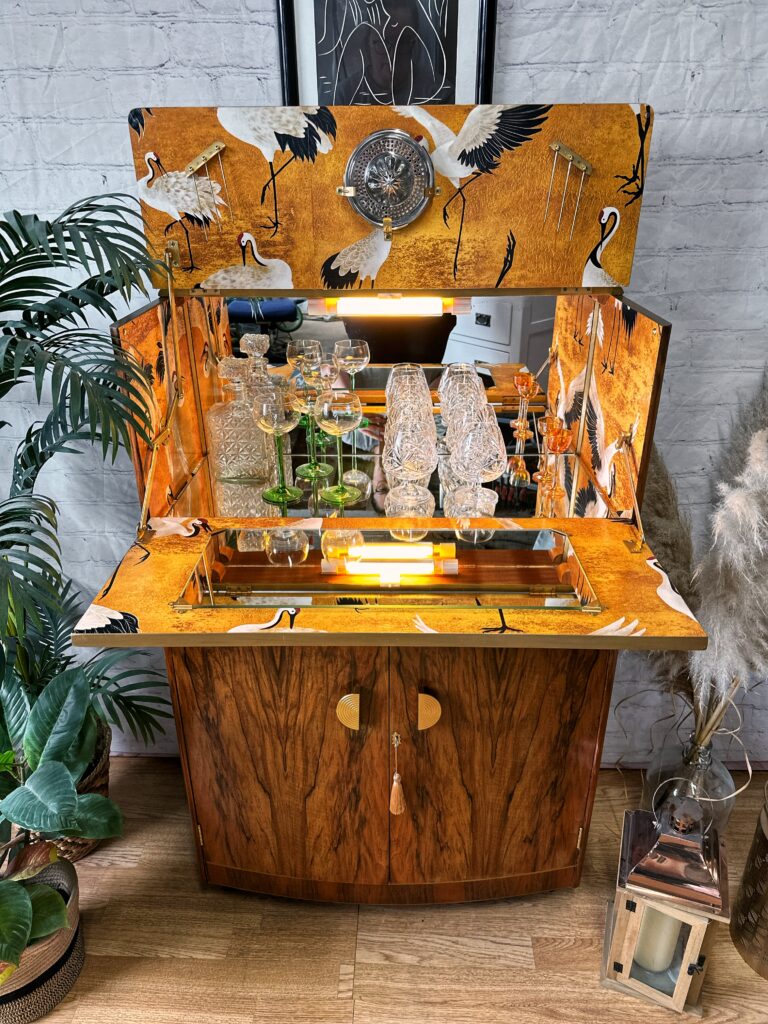 The Turnridge of London Cocktail Cabinet, a vintage walnut Art Deco piece from the 1950s, features a mirrored back and yellow crane-patterned wallpaper. The retro bar cabinet is adorned with glassware, decanters, and decorative bottles. Above the cabinet are displayed a tribal shield and spears. Nearby, plants and framed artwork enhance the stylish setup.