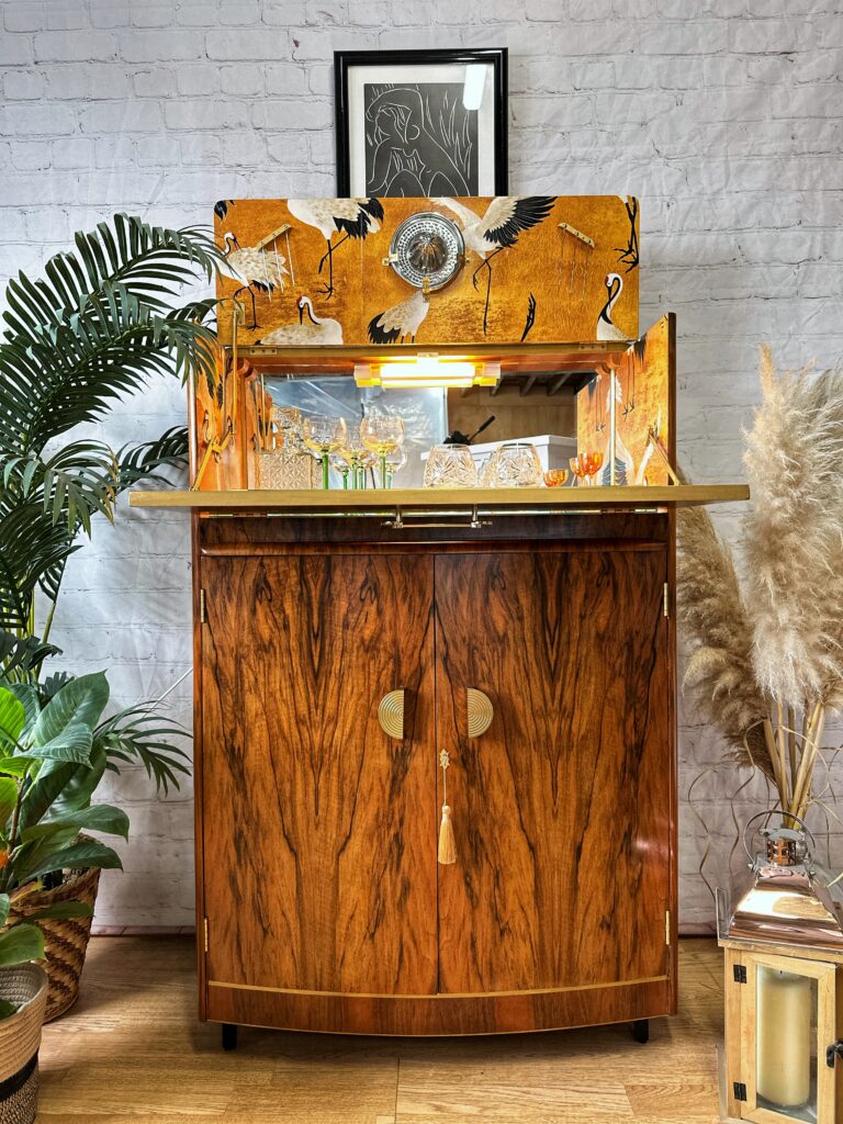 A stylish, vintage Turnridge of London Cocktail Cabinet from the 1950s with an open top displaying glassware and a decorative golden bird motif. It stands against a textured white brick wall, framed by green plants on the left and beige pampas grass on the right. A framed artwork hangs above.