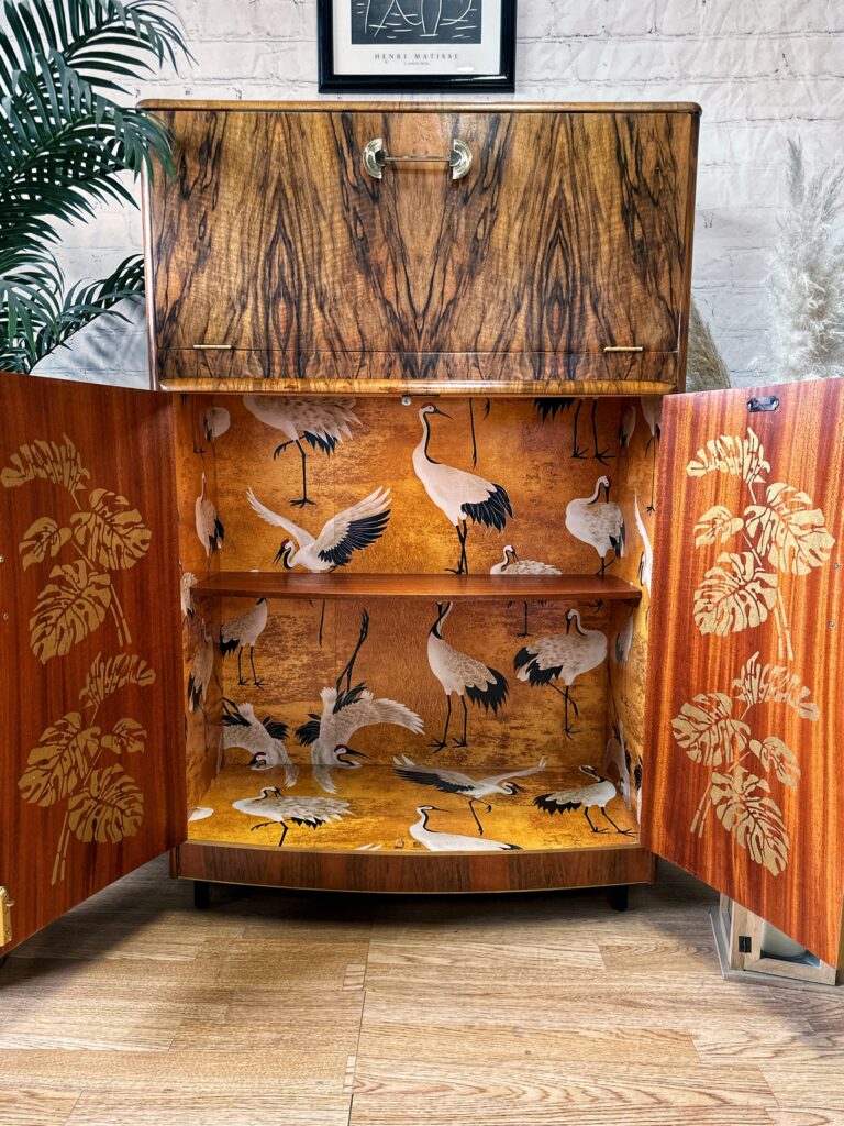 The Turnridge of London Cocktail Cabinet, a vintage 1950s Art Deco drinks bar made from walnut, stands open to reveal an interior adorned with crane and flower motifs. The exterior showcases a rich wood grain finish. It is set against a light-colored brick wall with a framed art piece above and flanked by potted plants.