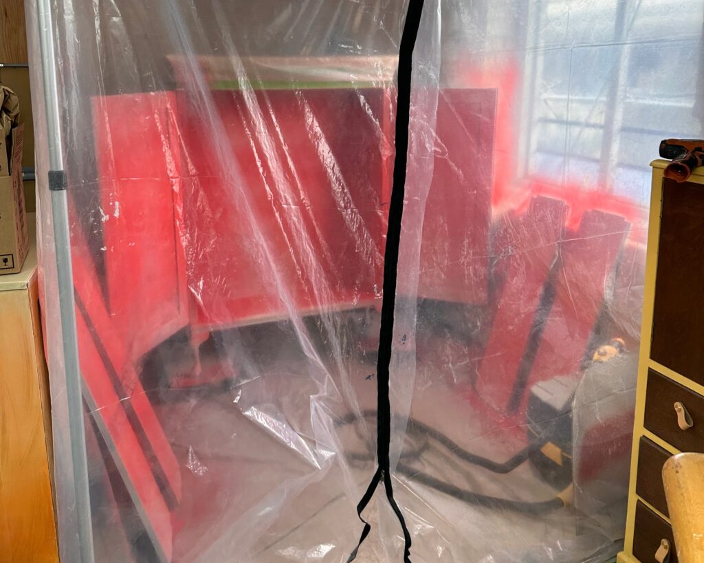 A workshop area is enclosed with a transparent plastic sheet, secured with a black zipper. Behind the plastic, there are several red-painted wooden boards and various workshop tools. The setup suggests a painting or construction project in progress.