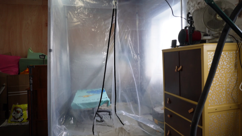 A room with walls covered in clear plastic sheets contains a blue table with a black object on it. The table sits slightly off-center, and there are various objects around the room, including a fan, a dresser, and boxes. Electrical cords hang from the ceiling.