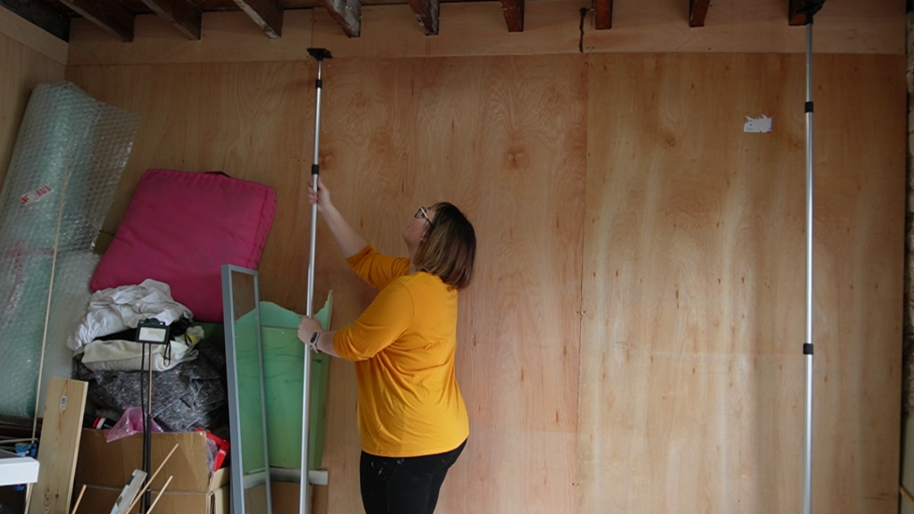 A woman wearing a yellow shirt is standing in a room with a wooden wall and ceiling. She is erecting a pole from the floor to the ceiling. To her left is a pile of miscellaneous items, including a pillow, a bubble-wrapped frame, and boxes.