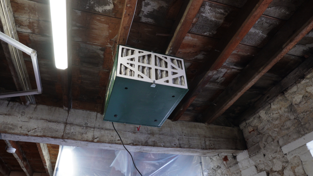 An air filter unit is mounted on the ceiling of a room with exposed wooden beams and a mix of concrete and stone walls. A fluorescent light fixture and plastic sheeting are visible nearby. The filter has a white grid pattern and a green base.