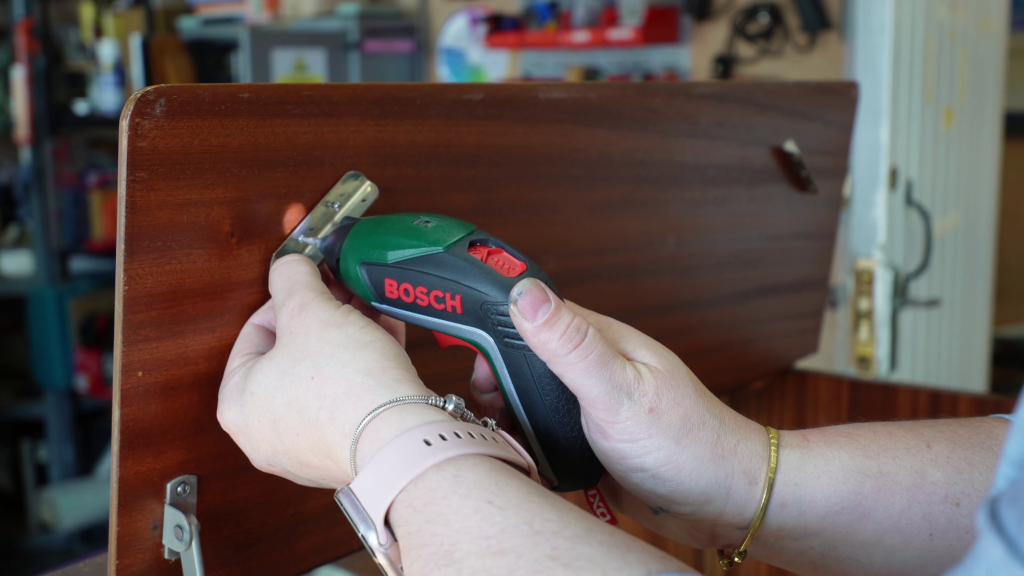 A person using a Bosch electric screwdriver to assemble or repair a wooden piece of furniture. The person's wrist is adorned with a smartwatch and a bracelet. Various tools and items are visible in the blurred background.