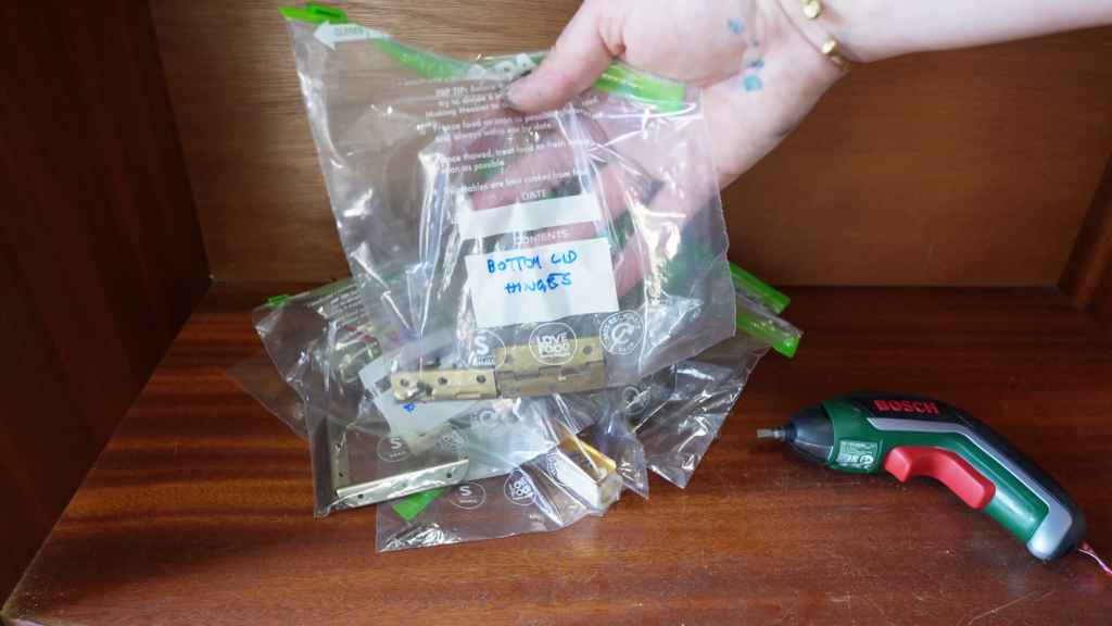 A tattooed hand holds a clear plastic bag labeled "Bottom Cup Hinges" containing metal hardware items. Several similar bags full of various hardware are laid out on a wooden surface. A green Bosch electric screwdriver lies next to the bags.