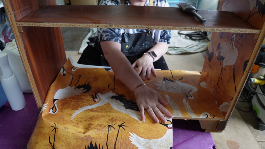 A person is seen applying wallpaper with white crane designs on a yellow background to a piece of wooden furniture. Their hands are smoothing the wallpaper as they work. A brush and some tools can be seen nearby.