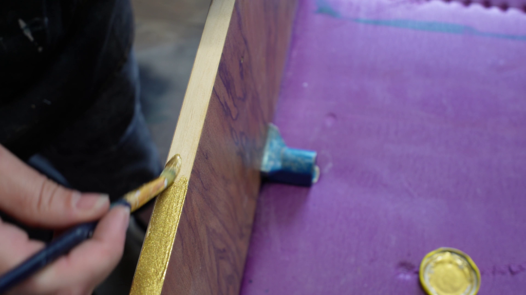 A person's hand holding a paintbrush is applying gold paint to the edges of a wooden surface. The wooden piece is placed on a purple fabric or material. A small blue clamp is partially visible, securing the wood in place.