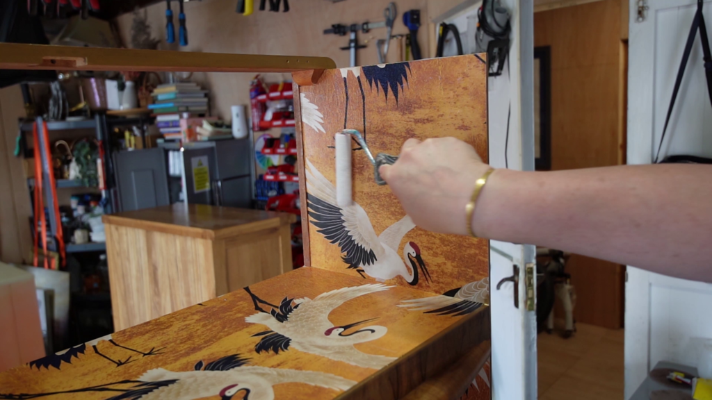 A person's hand, wearing a bracelet, is seen adjusting a metal handle on a wooden object with a painted design featuring cranes. The background shows a cluttered workshop with a wooden table, shelves, and various tools on the walls.