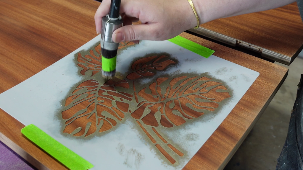 A close-up of a person's hand holding a small can of spray paint, filling in a stencil of a large leaf. The stencil is secured on a wooden surface with green tape. The painted areas on the stencil are brownish-orange.