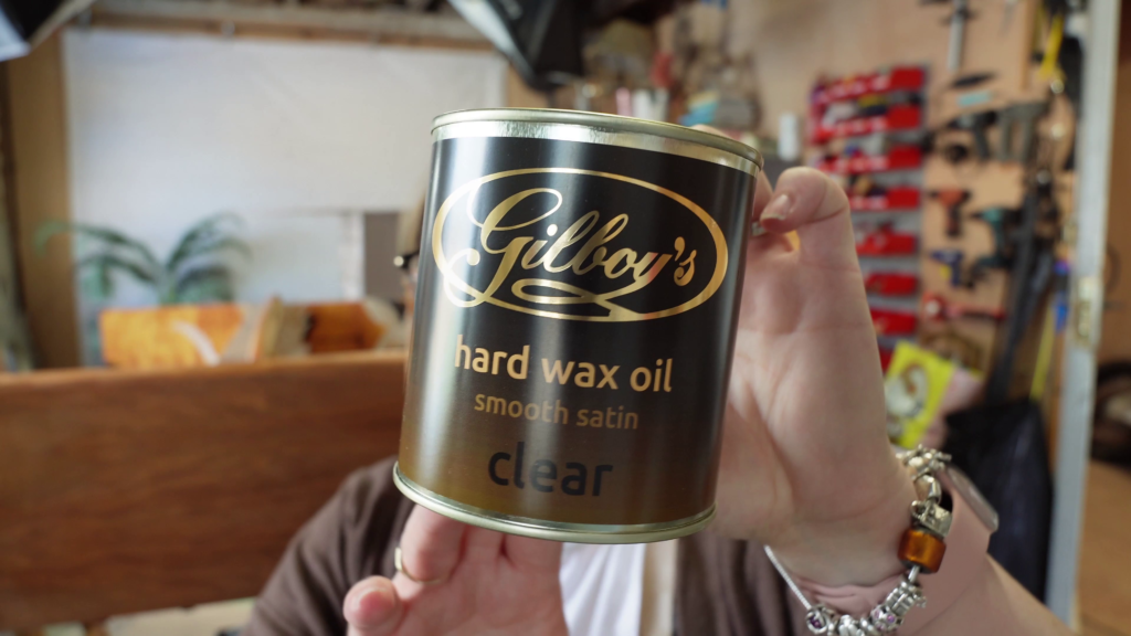 A person wearing a brown sweater is holding up a can of Gilboy's Hard Wax Oil in clear smooth satin finish. The background appears to be a workshop with tools and equipment. The person's face is not visible.