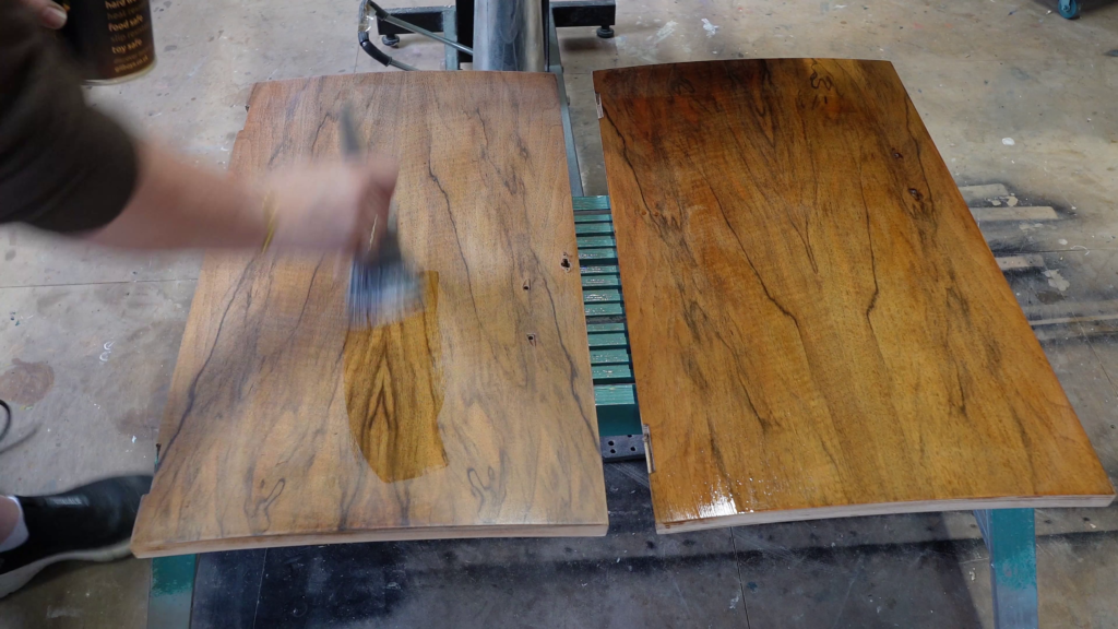 A person is applying varnish with a brush onto a wooden plank, which is beside another plank that has already been varnished. The varnished plank has a glossy finish, while the plank being worked on is still partially matte. The background is a workshop.