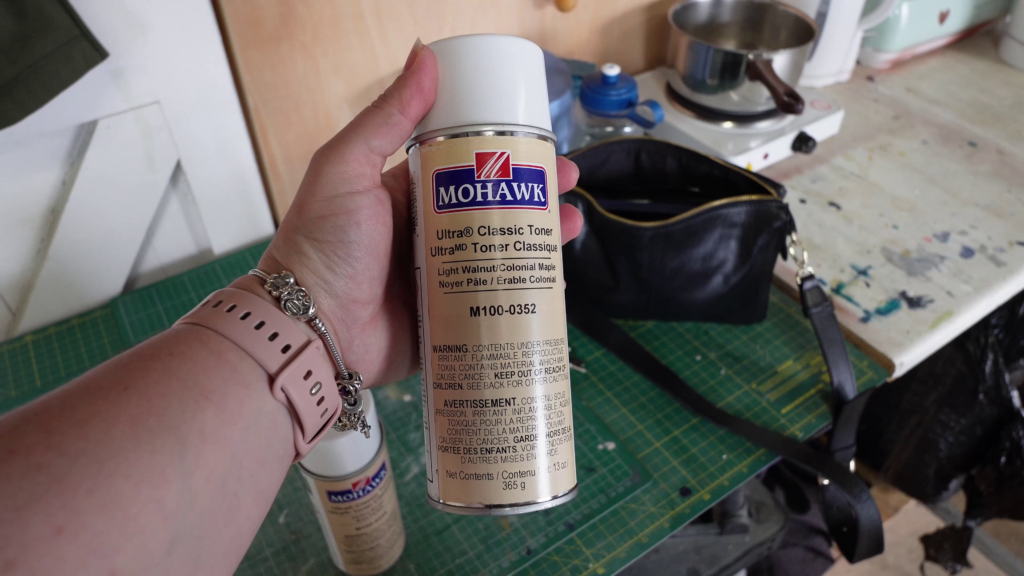 Close-up of a person holding a can of Mohawk Ultra® Classic Toner, labeled "M100-0352 Light Walnut / Colonial Maple." The person is wearing a pink watch and bracelet. A cutting mat, a black bag, and various items are visible in the background.