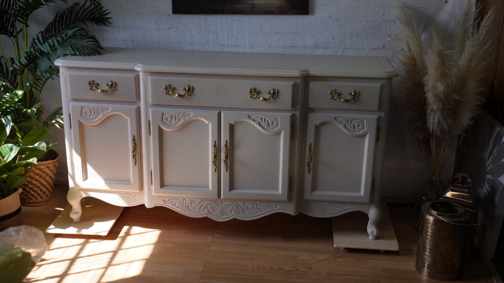 A white vintage sideboard with ornate gold handles and intricate carvings sits in a sunlit room with wooden flooring. Surrounding plants, including a potted fern and pampas grass, add a touch of greenery.