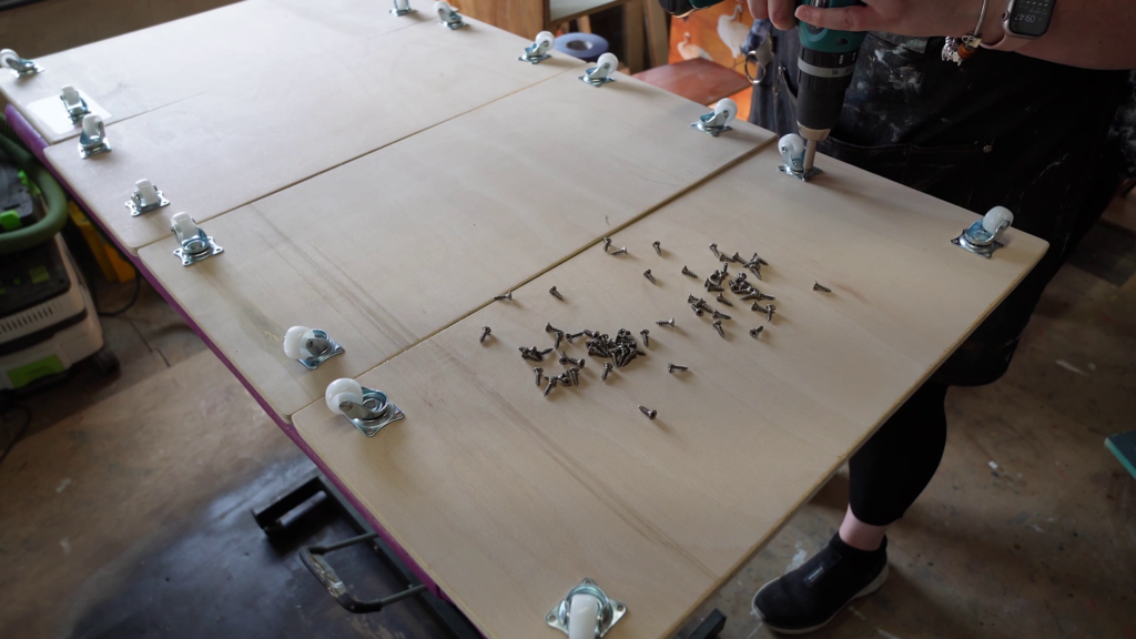 A person is using a power drill on three large wooden panels held together with clamps. Several caster wheels are attached to the panels' edges, and wood shavings are scattered on the surface. The person is wearing dark clothing and sneakers.