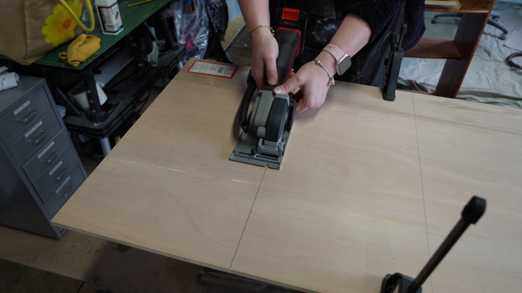 A person uses a circular saw to cut a piece of plywood in a workshop. They wear a watch and bracelet on their left wrist, and the workspace is filled with various tools and equipment. A red item with white labeling is also visible on the plywood.