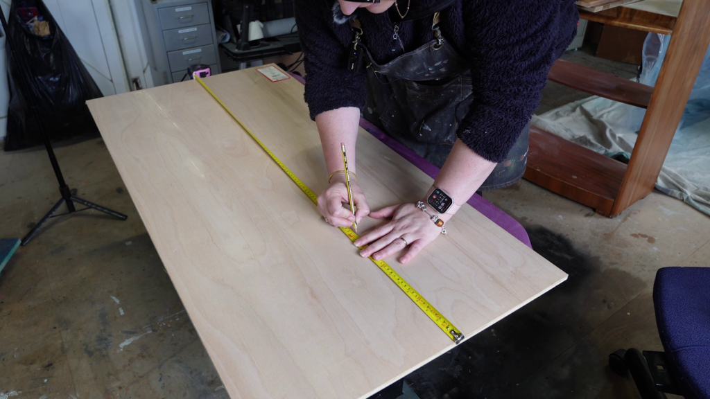 A person is marking a measurement on a piece of plywood using a pencil and measuring tape. The person is wearing a dark sweater, an apron, a smartwatch, and glasses, and they are working in a workshop setting with tools and equipment visible in the background.