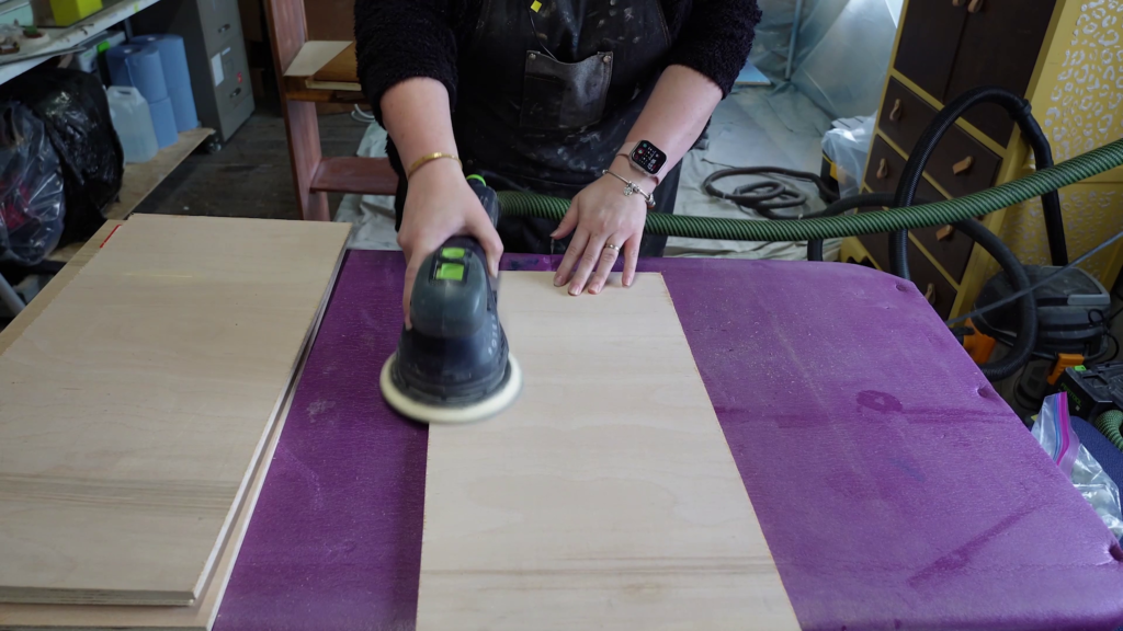 A person sands a piece of wood on a workbench using an electric sander. The workbench is covered with a purple cloth, and other woodworking tools are visible in the background. The person wears a black shirt and a smartwatch.