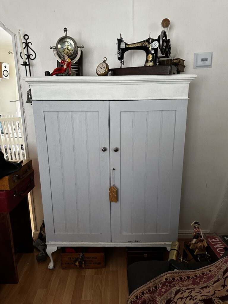 A white wooden cabinet with two doors stands in a room, adorned with vintage items on top, including an old sewing machine, a mantle clock, and other small decorations. There is a suitcase partially visible to the left and various other items around.