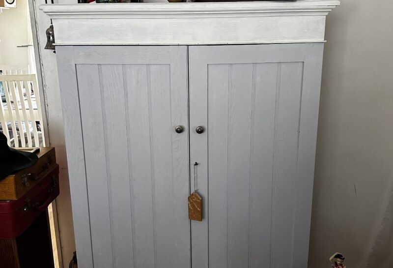 A white wooden cabinet with two doors stands in a room, adorned with vintage items on top, including an old sewing machine, a mantle clock, and other small decorations. There is a suitcase partially visible to the left and various other items around.