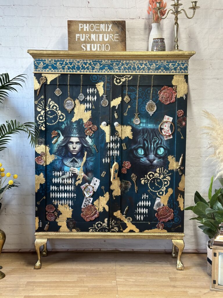 A wooden cabinet adorned with hand-painted Alice in Wonderland-themed art. There are images of Alice, playing cards, and the Cheshire Cat amidst a blue and black background with gold accents. It stands in a room with plants and a "Phoenix Furniture Studio" sign on top.