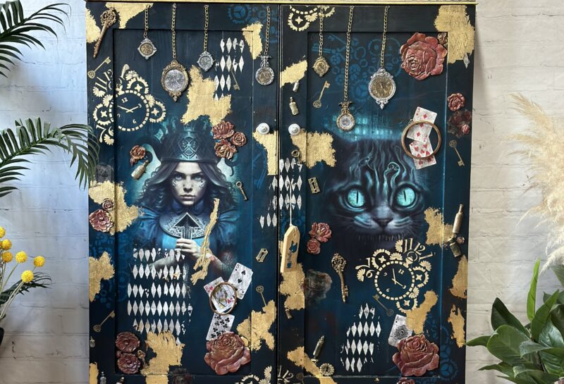 A wooden cabinet adorned with hand-painted Alice in Wonderland-themed art. There are images of Alice, playing cards, and the Cheshire Cat amidst a blue and black background with gold accents. It stands in a room with plants and a "Phoenix Furniture Studio" sign on top.