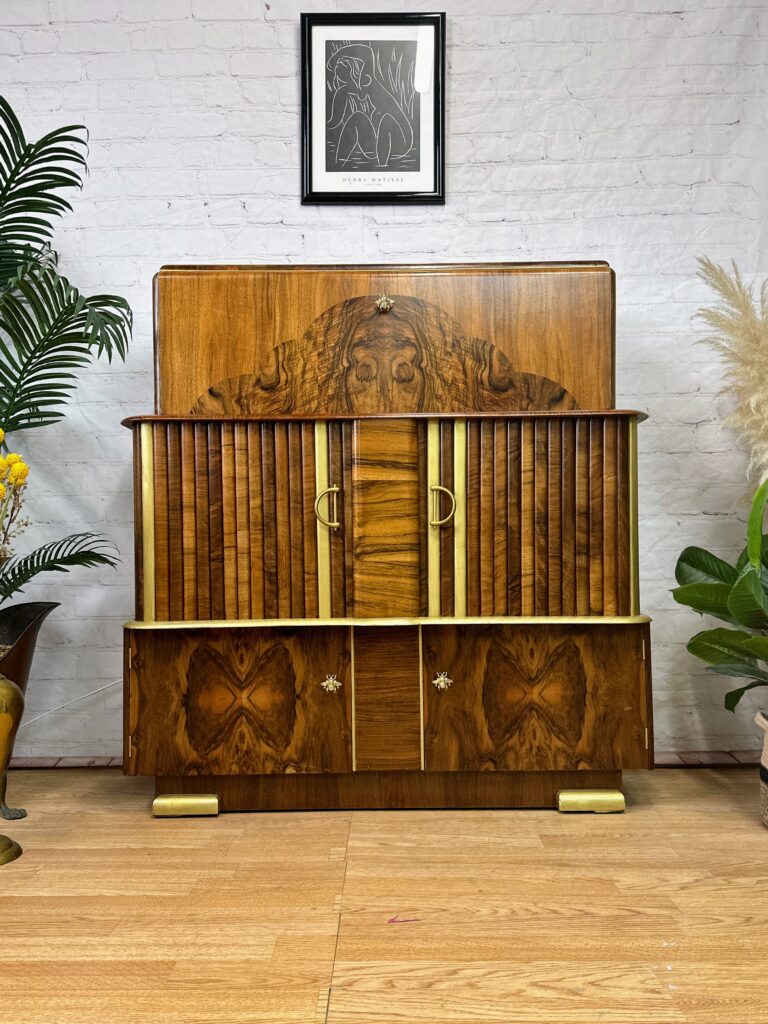 A wooden sideboard with intricate details stands against a white brick wall. The sideboard features a mix of vertical and horizontal patterns with two golden handles at the center. A framed artwork hangs above it, and plants are placed on either side.