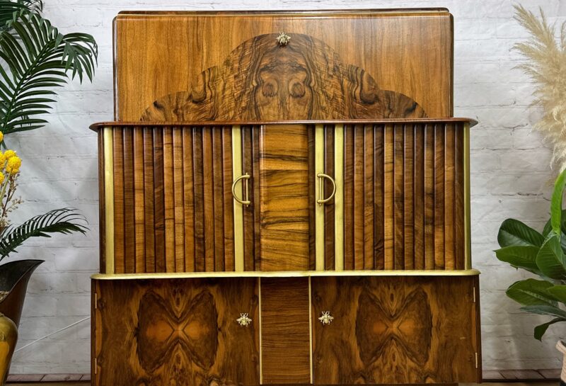 A wooden sideboard with intricate details stands against a white brick wall. The sideboard features a mix of vertical and horizontal patterns with two golden handles at the center. A framed artwork hangs above it, and plants are placed on either side.