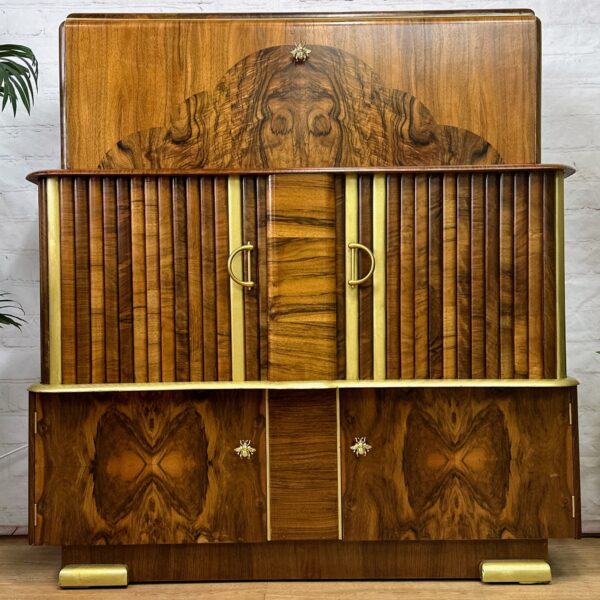 A vintage wooden cabinet with intricately designed wood grain patterns. The cabinet has two central doors with gold handles, flanked by vertical slats. Below, it features two smaller compartments with star-shaped knobs. Surrounding plants add a touch of greenery.