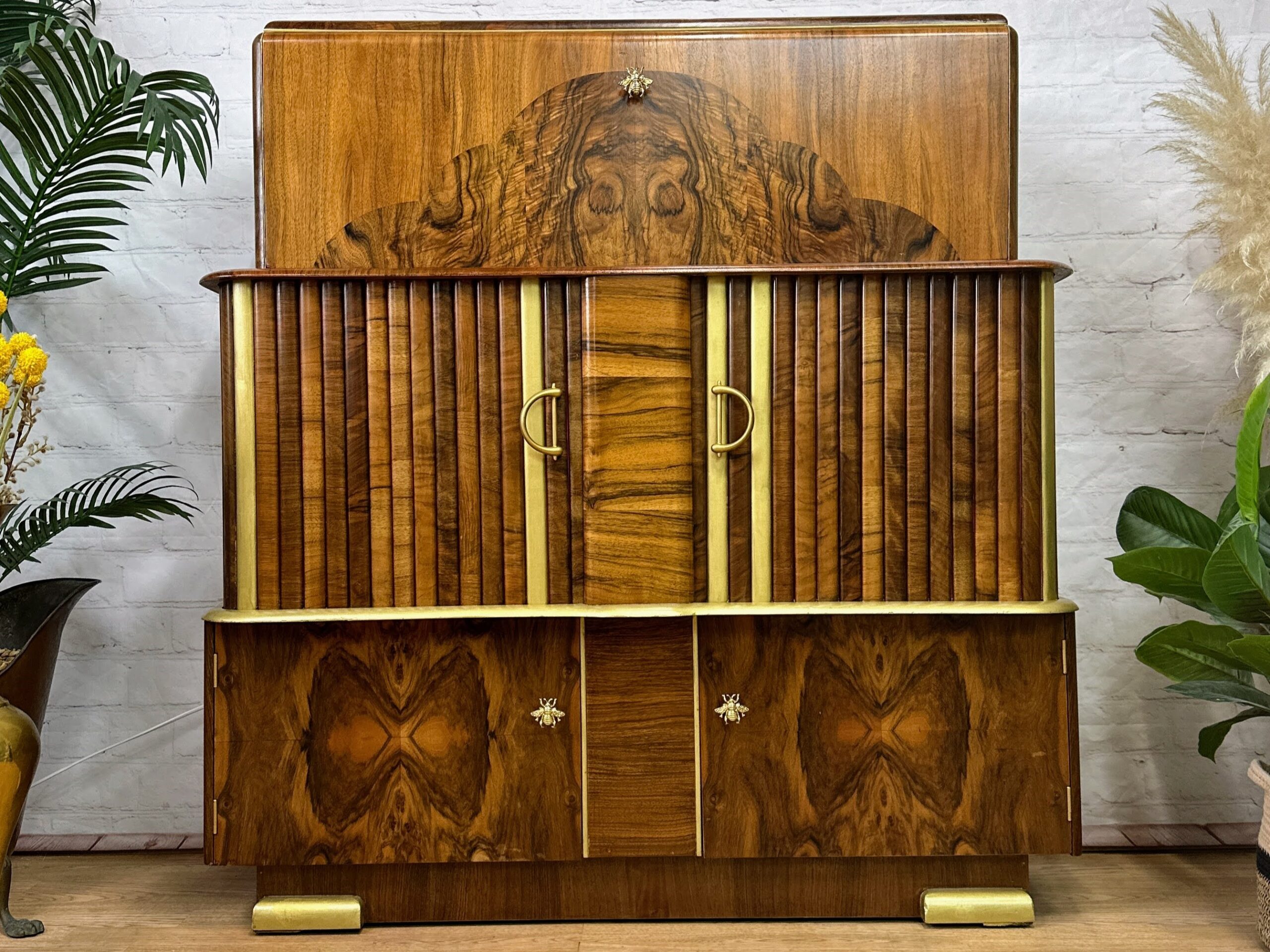 A vintage wooden cabinet with intricately designed wood grain patterns. The cabinet has two central doors with gold handles, flanked by vertical slats. Below, it features two smaller compartments with star-shaped knobs. Surrounding plants add a touch of greenery.