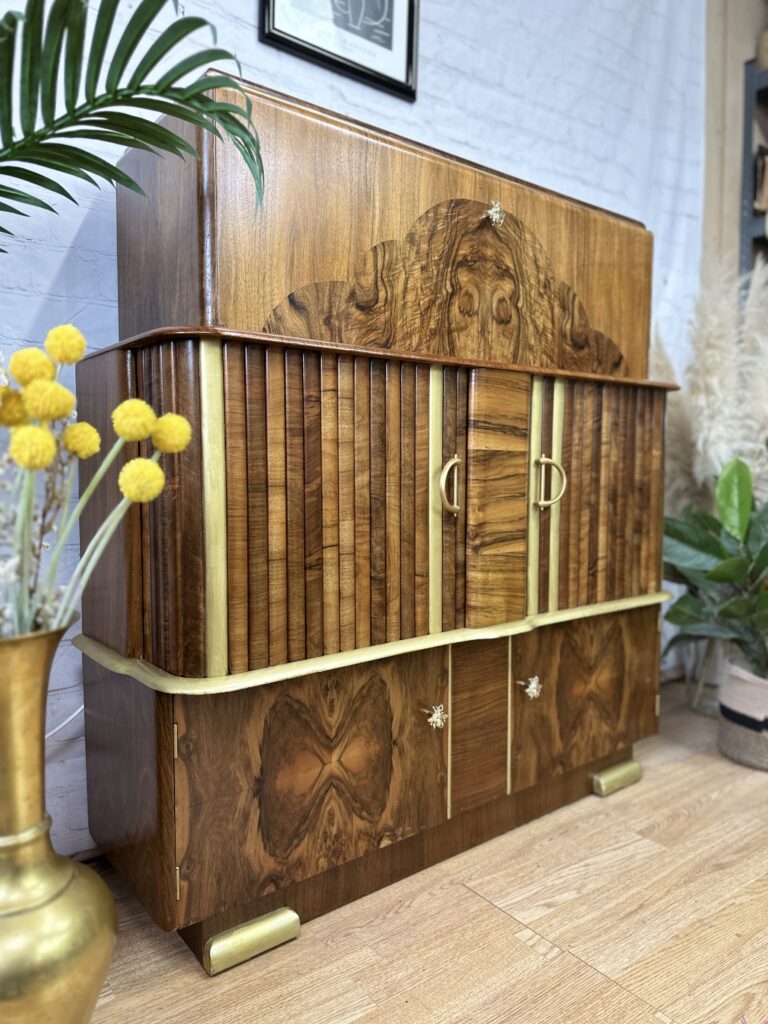 A vintage wooden cabinet featuring intricate wood inlay designs and brass accents stands on a hardwood floor. The cabinet has multiple compartments with central doors and a top section with a curved design. It is flanked by indoor plants and decorative yellow flowers in a brass vase.