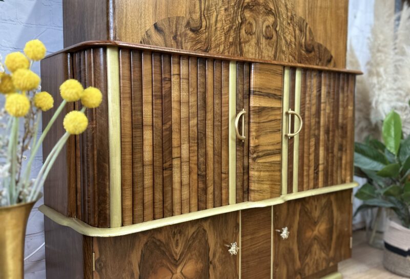 A vintage wooden cabinet featuring intricate wood inlay designs and brass accents stands on a hardwood floor. The cabinet has multiple compartments with central doors and a top section with a curved design. It is flanked by indoor plants and decorative yellow flowers in a brass vase.