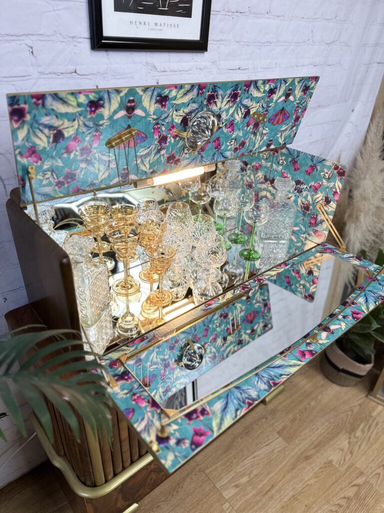 A vintage wooden bar cabinet is open, revealing an array of crystal and glassware. The interior is lined with a colorful floral pattern. Glasses of various shapes and sizes are displayed neatly inside. The background features a white brick wall and framed artwork.