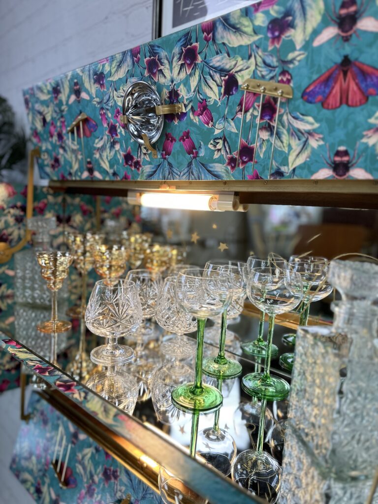 A vintage walnut cocktail cabinet, designed in the elegant Art Deco style of the 1930s, showcases an assortment of intricate crystal glassware. The Retro Bar features floral and butterfly designs, and the glassware includes detailed goblets with green stems and etched patterns, all reflecting the light beautifully.