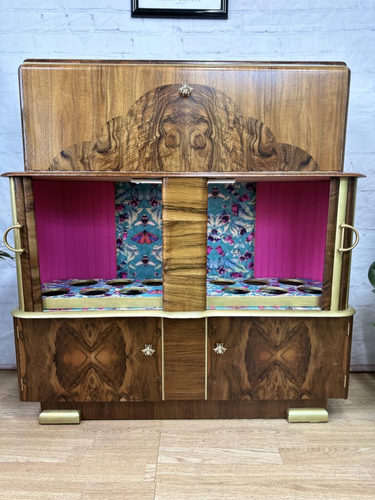 An Art Deco walnut cocktail cabinet, featuring ornate grain patterns and brass handles, stands against a white brick wall. The interior shelves are lined with vibrant pink floral fabric. This vintage drinks unit from the 1930s boasts two doors and offers a retro bar style that exudes mid-century modern charm.