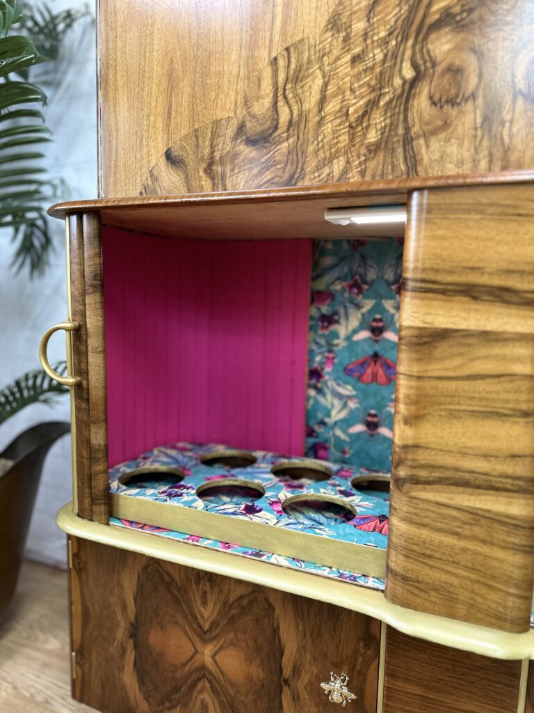 A wooden cabinet with an open section displaying a colorful interior. The interior has a vibrant pink back panel and a blue, butterfly-patterned floor with circular cutouts. The cabinet also features handles and a decorative bee emblem on the lower right door.