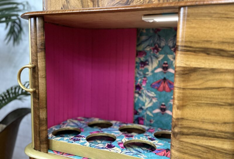 A wooden cabinet with an open section displaying a colorful interior. The interior has a vibrant pink back panel and a blue, butterfly-patterned floor with circular cutouts. The cabinet also features handles and a decorative bee emblem on the lower right door.