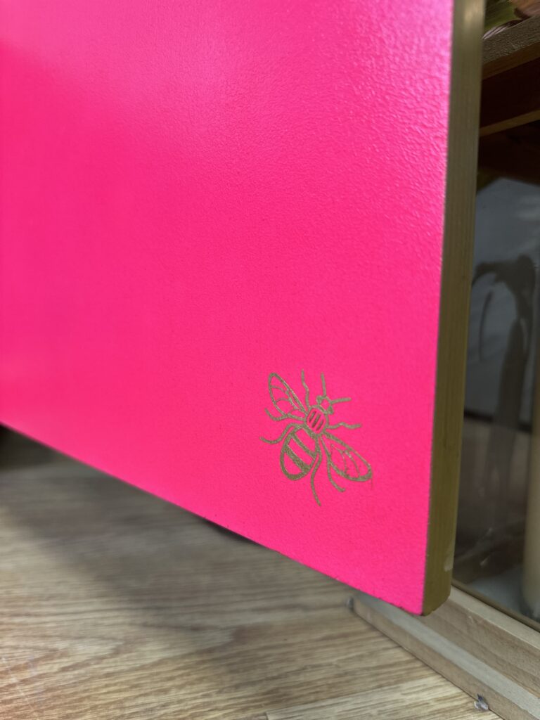 A close-up shot of a bright pink box edge with a small golden bee illustration in the bottom right corner. The box is placed on a wooden surface, and the edge of another glass-fronted box is visible in the background.