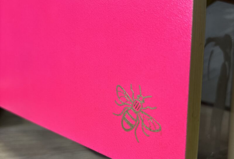 A close-up shot of a bright pink box edge with a small golden bee illustration in the bottom right corner. The box is placed on a wooden surface, and the edge of another glass-fronted box is visible in the background.