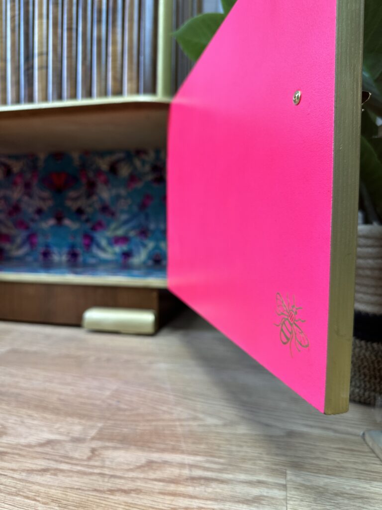 A vintage 1930s Art Deco walnut cocktail cabinet with a partially open door reveals a bright pink interior adorned with a bee illustration on the inner side of the door. The background displays a patterned, vibrant-colored interior, and a plant leaf is visible in the upper right corner.