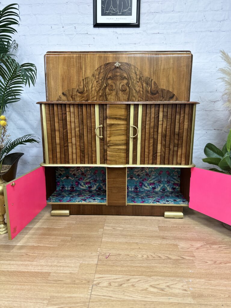 A vintage wooden cabinet with two open doors revealing a colorful, floral patterned interior. The cabinet has a detailed wood grain design on the front, brass handles, and stands on short brass-tipped legs. It is set against a light brick wall near a plant.