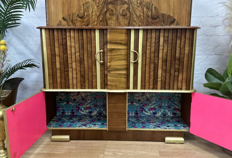 A vintage wooden cabinet with two open doors revealing a colorful, floral patterned interior. The cabinet has a detailed wood grain design on the front, brass handles, and stands on short brass-tipped legs. It is set against a light brick wall near a plant.